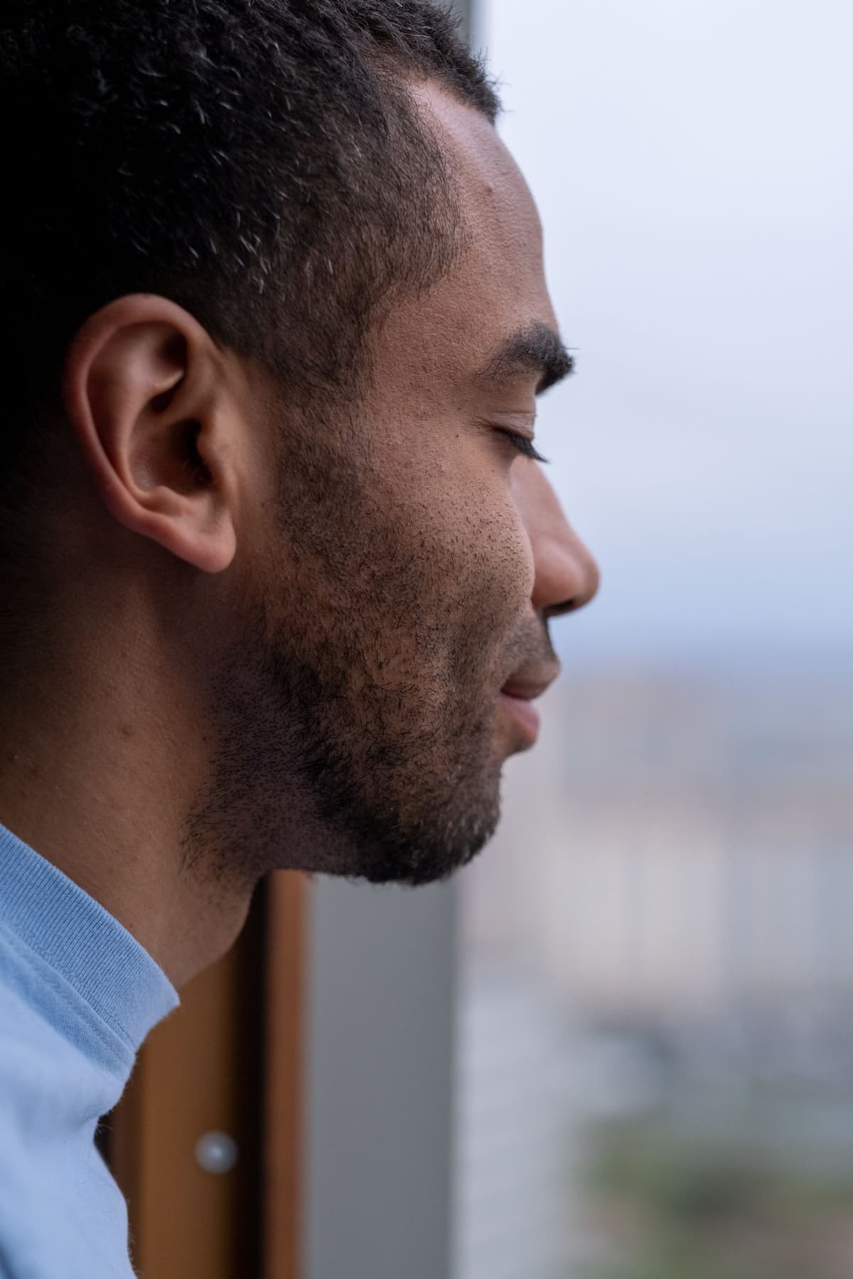 A person pausing with closed eyes beside a window in a quiet moment.