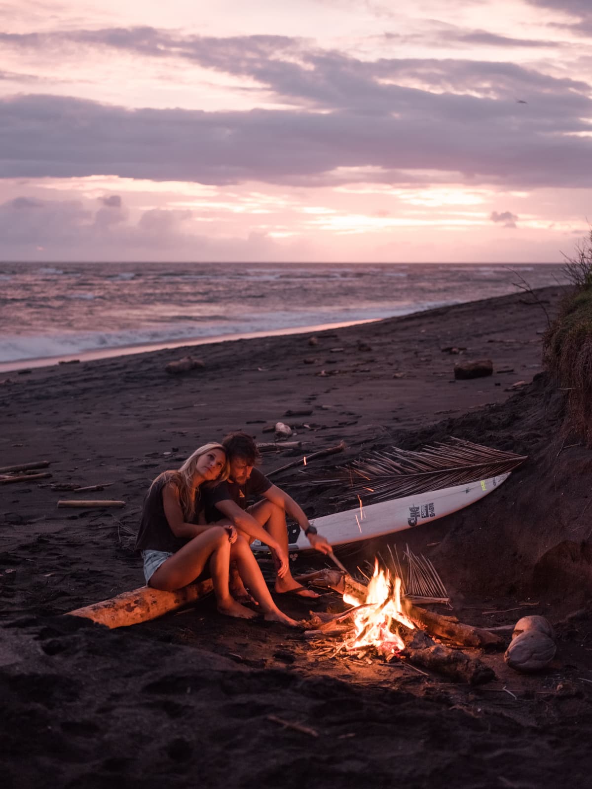 A couple sitting together by a beach fire at dusk.