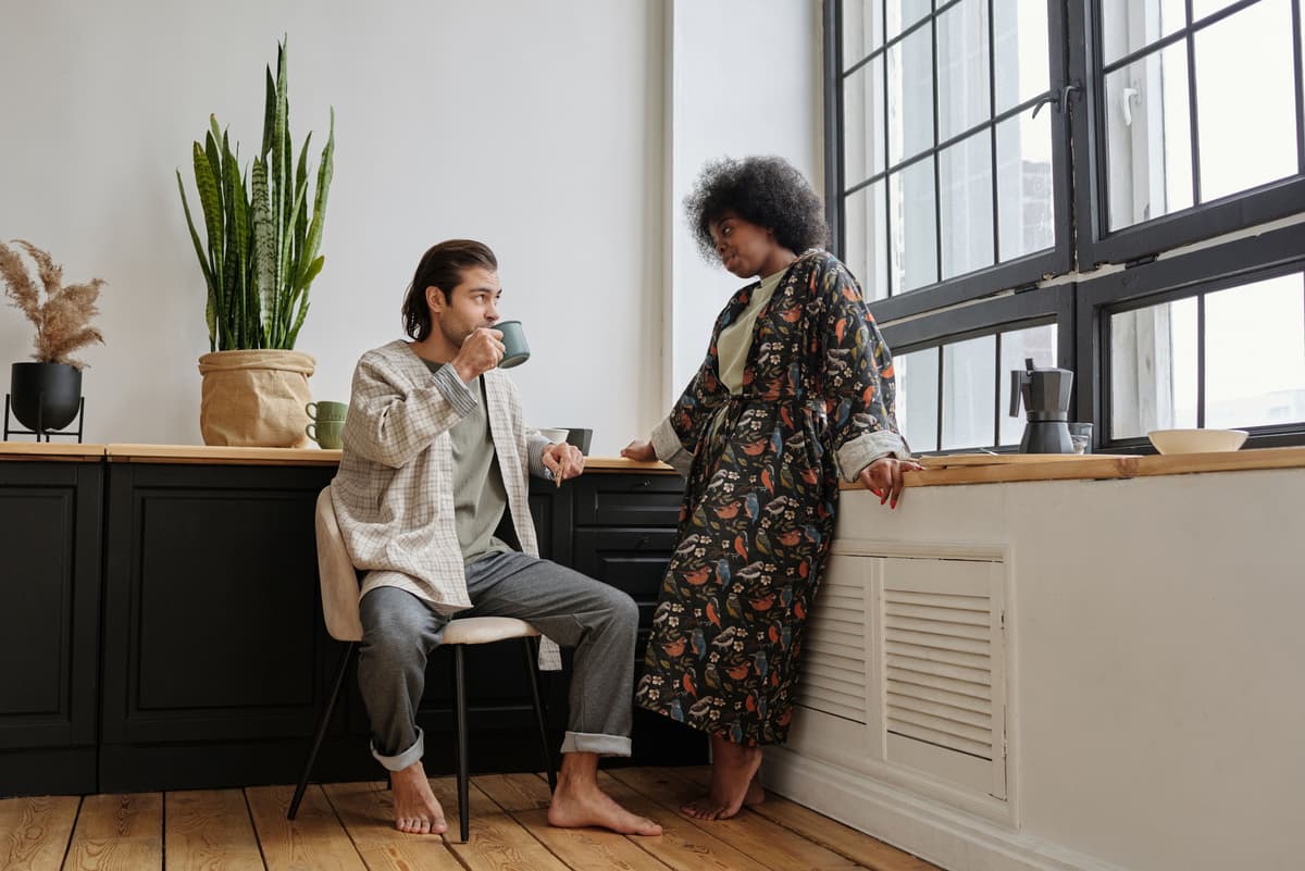 Two people talking over mugs near a window.