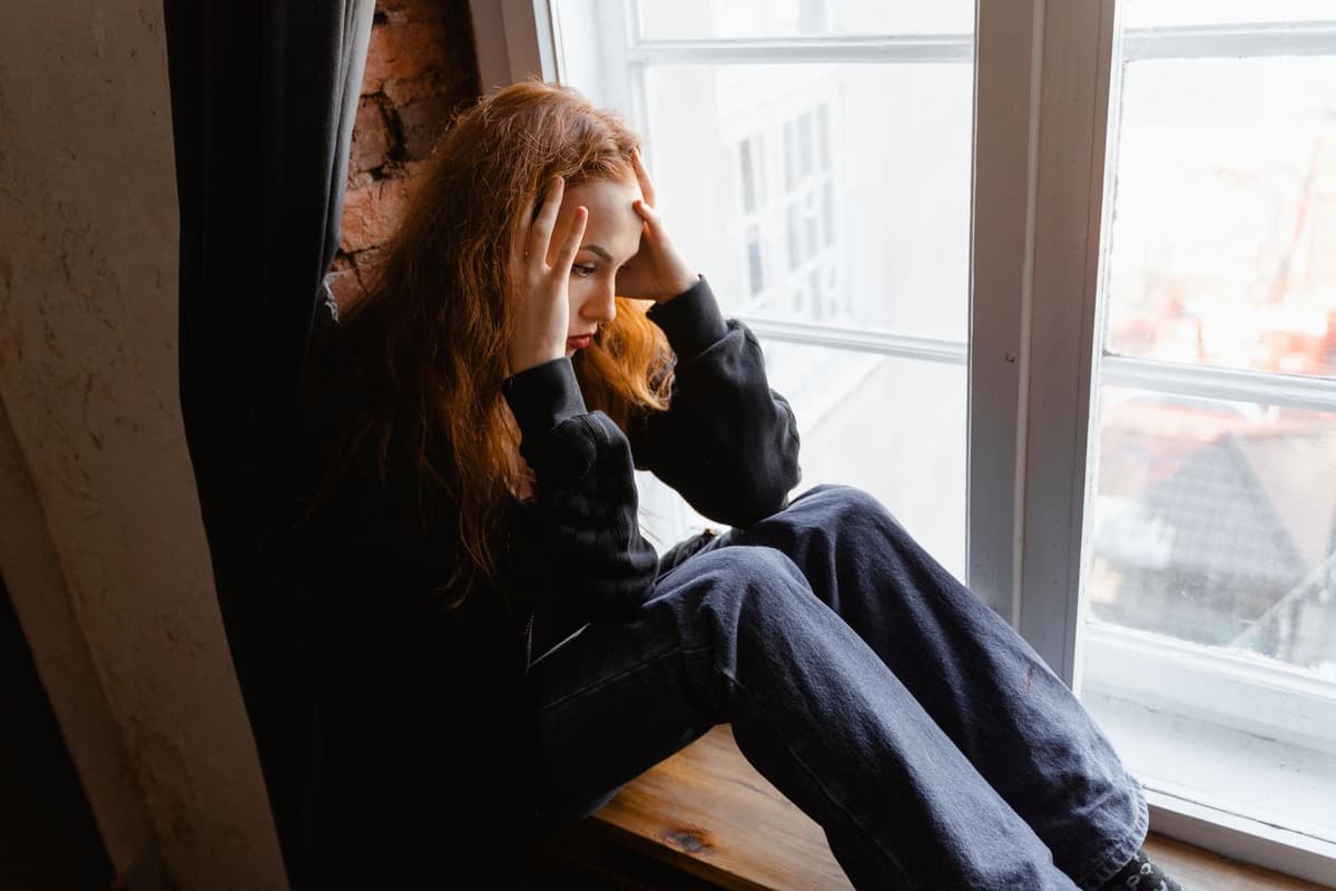 A person sitting alone by a window with their hand on their forehead.