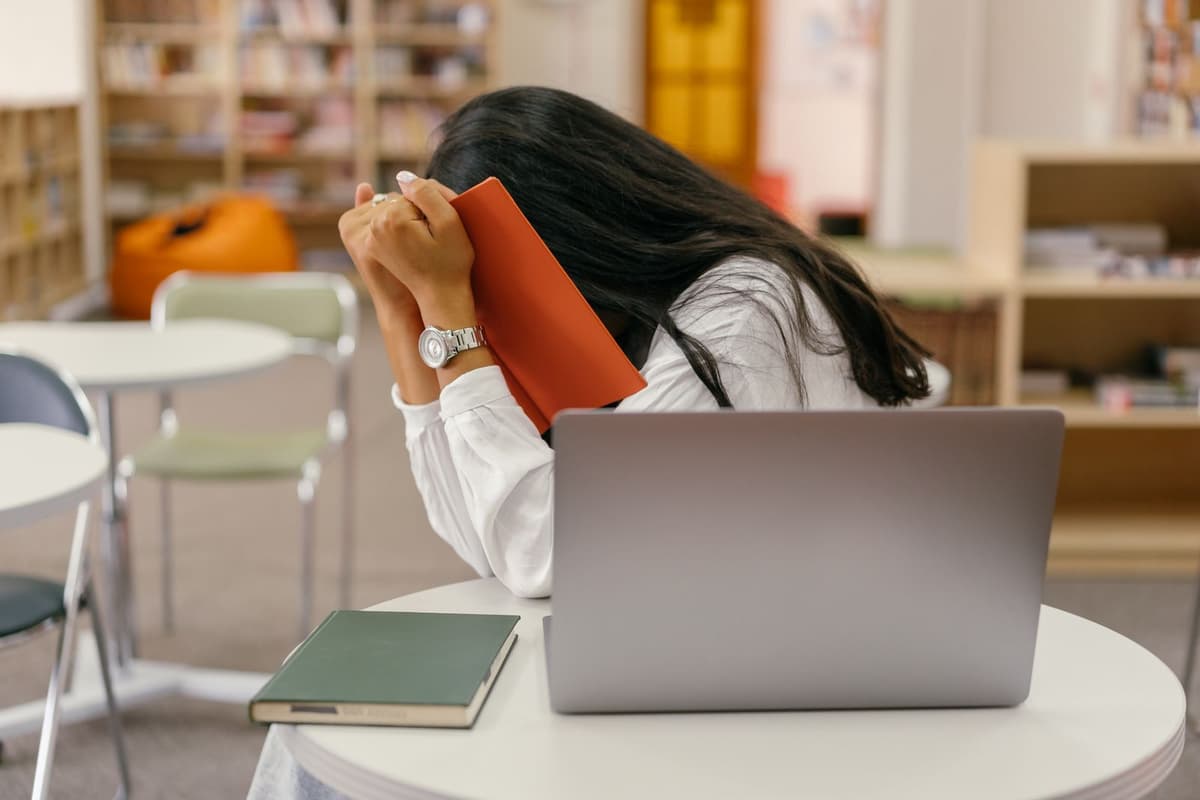 A person covering their face with a notebook while seated at a desk.